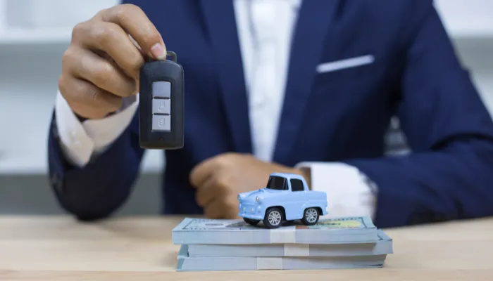 Homem segurando a chave de um carro, com carro em miniatura em cima de bolos de dinheiro, em sua mesa de escritório.